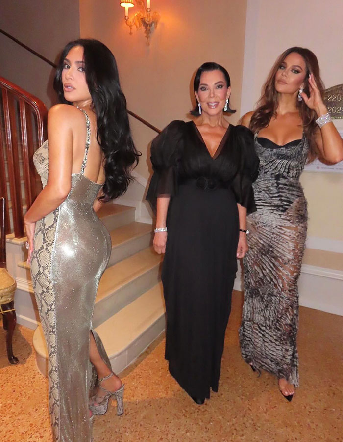 Kim Kardashian stuns in shimmering lingerie outfit, posing with two women in elegant dresses at a stylish indoor event.
