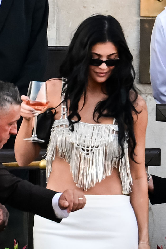 Woman in a revealing fringe top and white skirt at a racy pajama-themed afterparty holding a drink and wearing sunglasses.