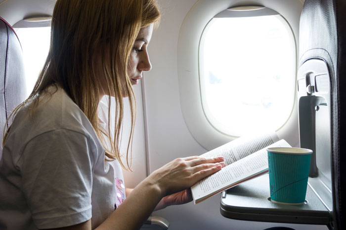 Passenger reading a book by the airplane window with a cup on the tray table during a flight.