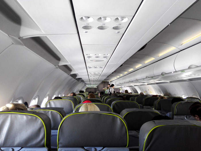 Inside an airplane cabin with rows of passengers seated, a flight attendant stands in the aisle addressing a situation.