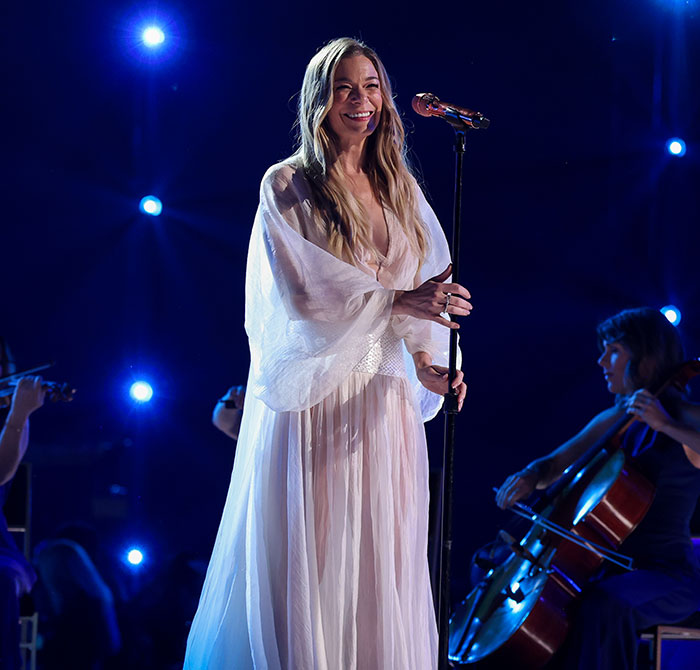 LeAnn Rimes performing on stage in a flowing white dress with musicians playing string instruments nearby. LeAnn Rimes performing on stage in a flowing white dress with musicians playing string instruments nearby.