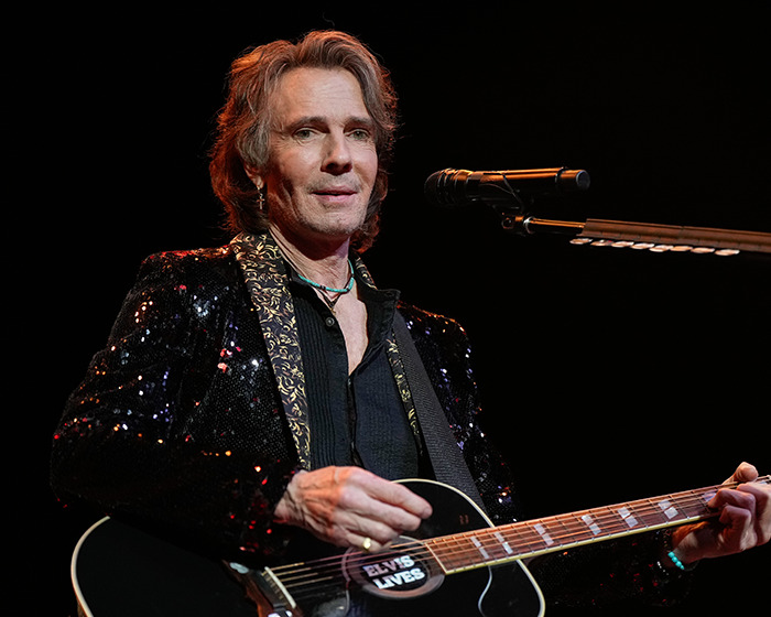Famous 80s rockstar performing on stage with guitar, wearing a black glittery jacket under dramatic lighting.