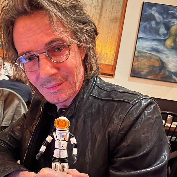 Famous 80s rockstar wearing glasses and leather jacket, holding an astronaut figure in a casual indoor setting.