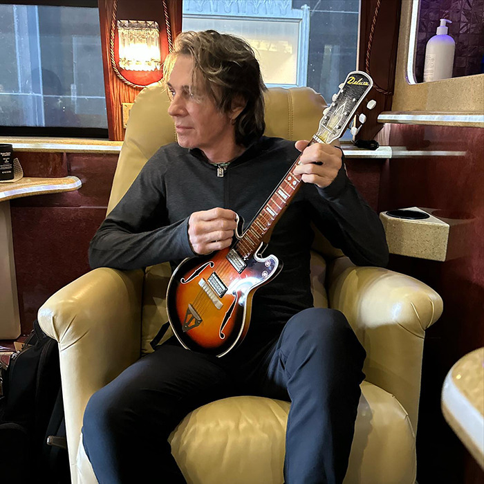 Famous 80s rockstar at 75 playing guitar while sitting in a cozy chair inside a tour bus or lounge area.