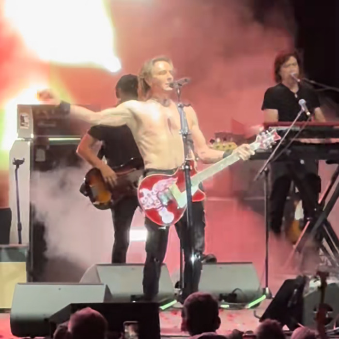 Shirtless famous '80s rockstar performs with guitar on stage, stunning the audience with his energetic live show.