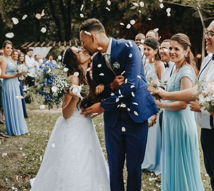 A millennial couple kissing at a wedding ceremony with guests throwing flower petals outdoors.