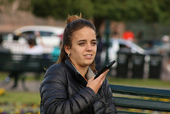 Young woman sitting on a bench outdoors, speaking into her smartphone, symbolizing longtime friends and ghost friend birthday calls.