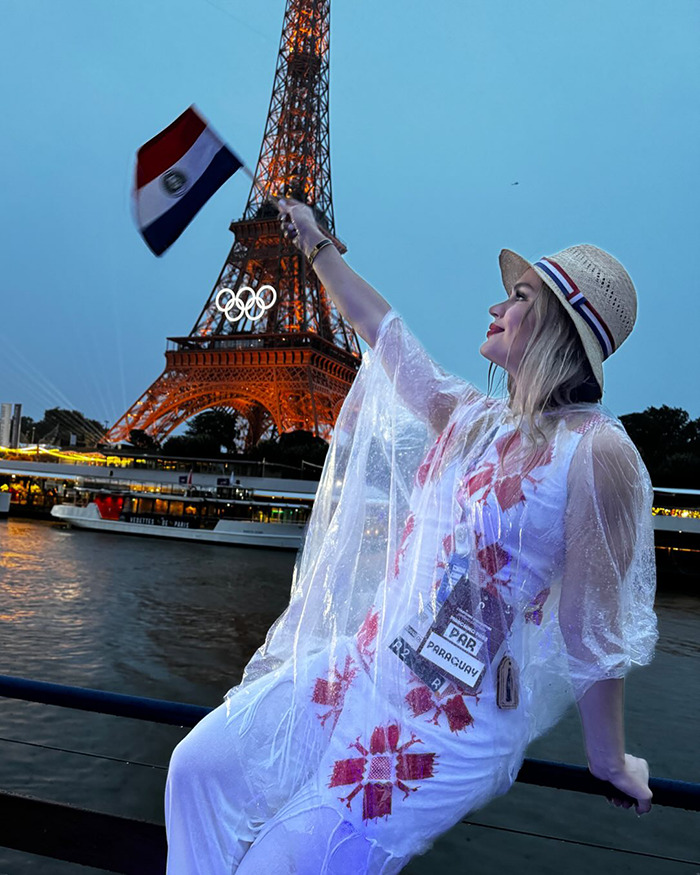 Woman in festive outfit waving Paraguayan flag near Eiffel Tower with Olympic rings, related to swimmer kicked out of Olympic Village. Woman in festive outfit waving Paraguayan flag near Eiffel Tower with Olympic rings, related to swimmer kicked out of Olympic Village.