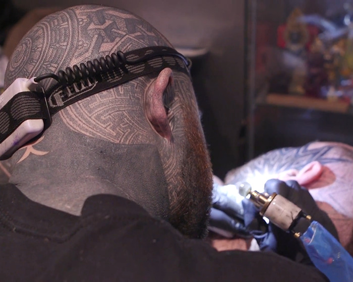 Man with face tattoos receiving tattoo work, showing detailed ink designs under focused lighting and wearing a headlamp. Man with face tattoos receiving tattoo work, showing detailed ink designs under focused lighting and wearing a headlamp.