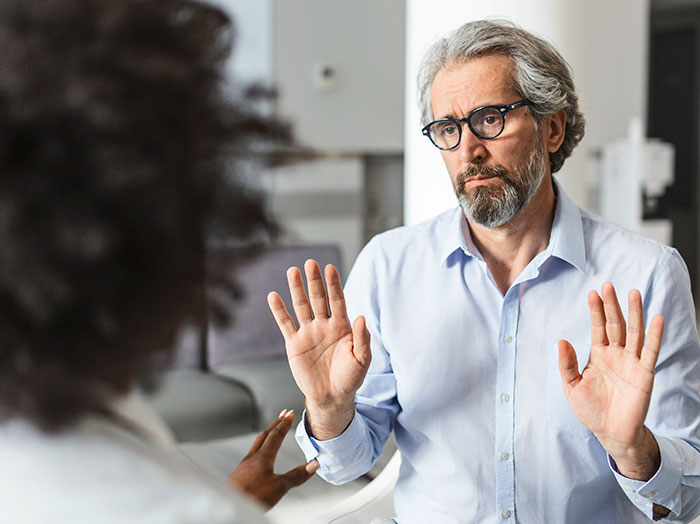 A man in glasses and a blue shirt raising hands during a serious conversation standing out in a rom-com moment.