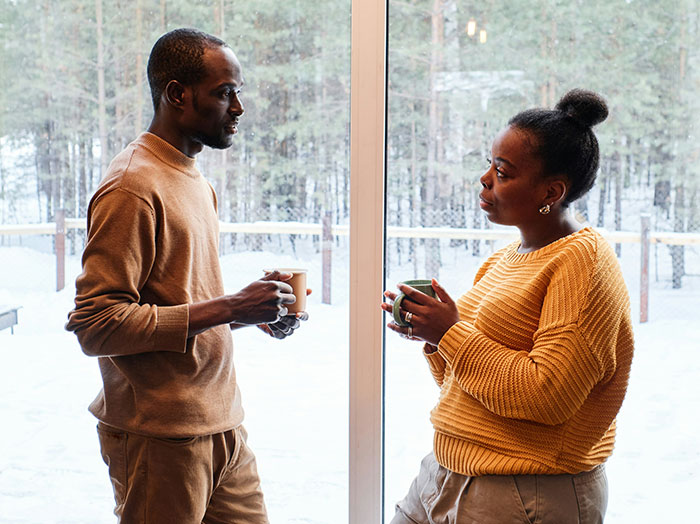 Man and woman holding mugs indoors by window, sharing a moment that makes him stand out in a romantic way