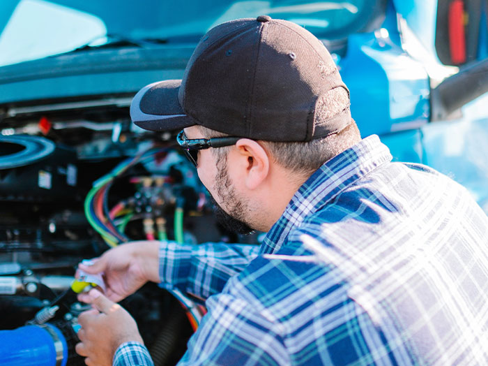 Man in a black cap and plaid shirt working on car engine, standing out with thoughtful and focused demeanor.