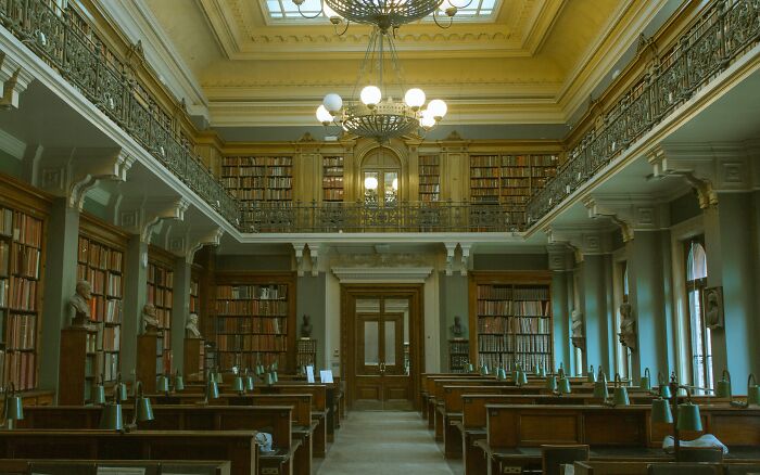 Grandiose library interior with wooden shelves, classic chandeliers, bust statues, and a skylight ceiling in a historic setting.