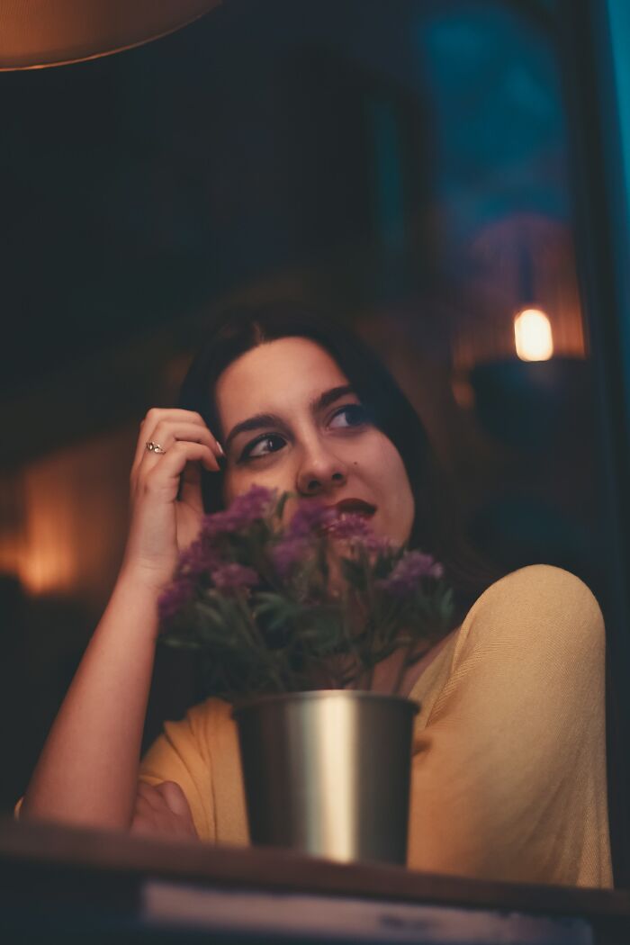 Young woman sitting thoughtfully indoors near a window with flowers, symbolizing hooking up with a friend’s parent experience.