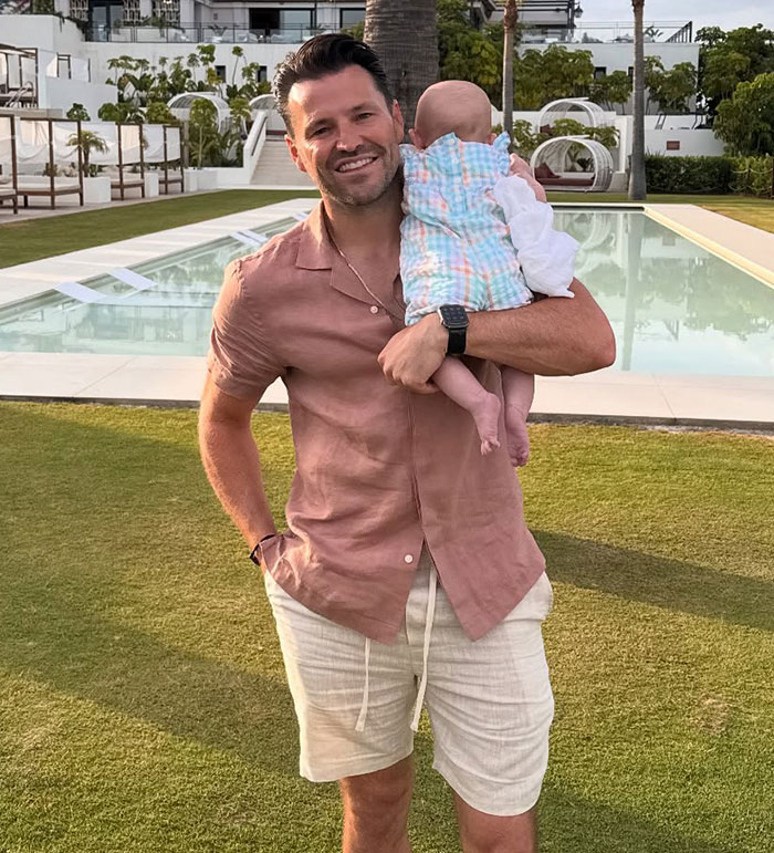 Man in casual summer clothes holding a baby outdoors near a pool, reflecting on stars forced to apologize for insensitive posts.