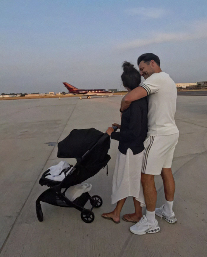 Couple embracing on an airport tarmac near a stroller with a small plane in the background after Air India tragedy.