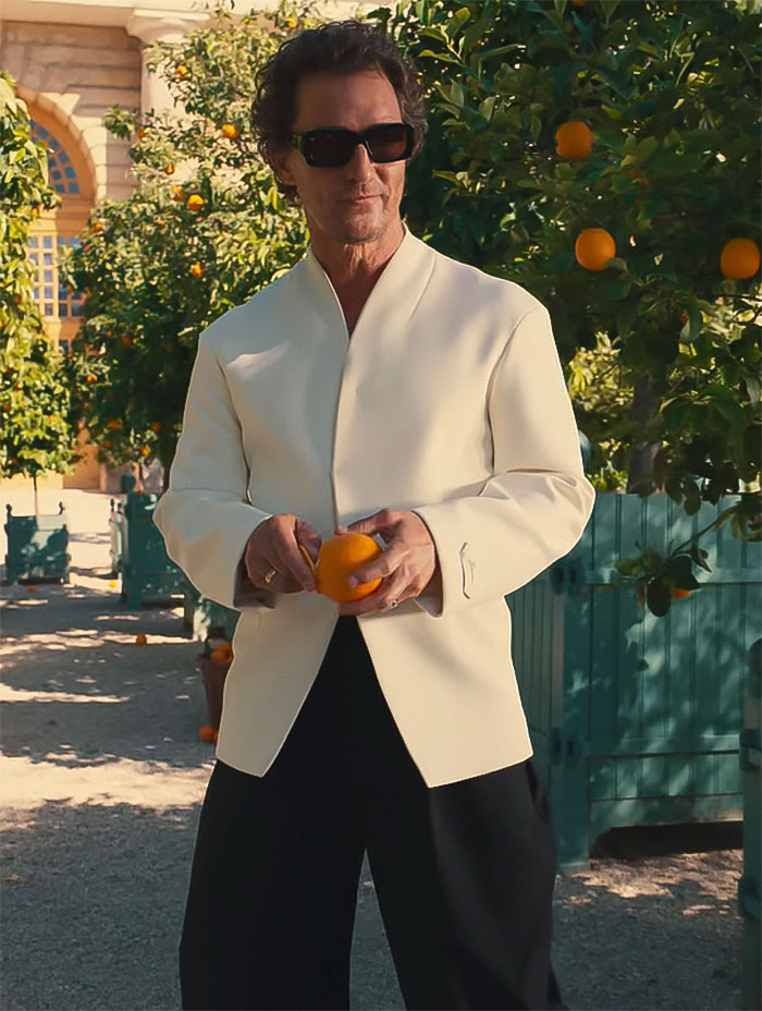 Matthew McConaughey wearing sunglasses and a white jacket holding an orange in a garden with orange trees around. Matthew McConaughey wearing sunglasses and a white jacket holding an orange in a garden with orange trees around.