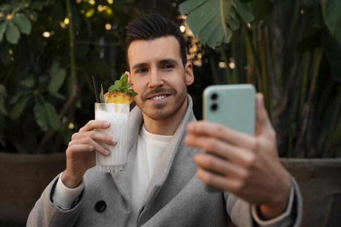 Man in a gray coat holding a cocktail and taking a selfie at a bar, illustrating dates that went sour moments.