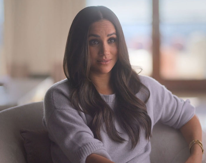 Meghan Markle sitting on a couch in a cozy sweater, sharing a controversial Hitchen hack in a bright room.