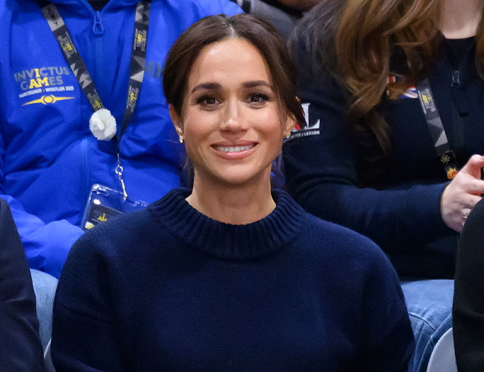 Meghan Markle smiling in a navy sweater at an event, related to Harry and Meghan's insufferable interview roast.