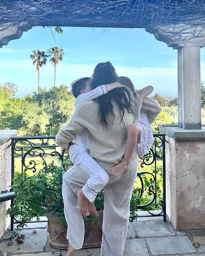 Meghan Markle carrying her kids outdoors on a sunny day with greenery and palm trees in the background.