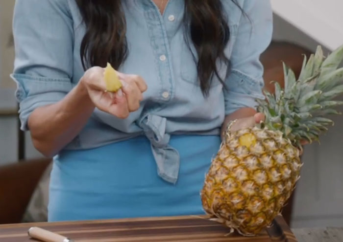 Woman demonstrating a kitchen hack by peeling a pineapple while wearing a blue shirt and apron in a home kitchen.