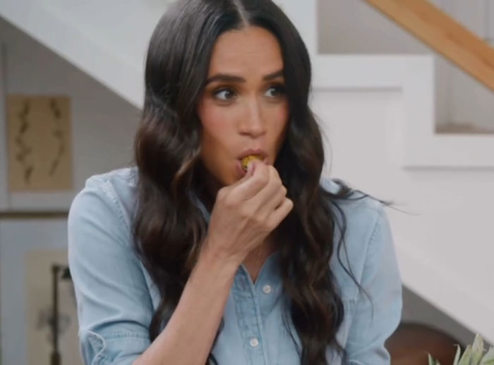 Meghan Markle eating a snack indoors, wearing a blue shirt, sharing a kitchen hack in a casual home setting.