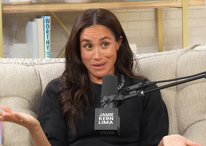 Meghan Markle speaking during an interview on the Jamie Kern Lima show with a playful expression on a beige couch.