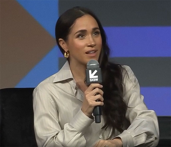 Meghan Markle speaking at SXSW event, holding microphone, wearing beige shirt and gold earrings.