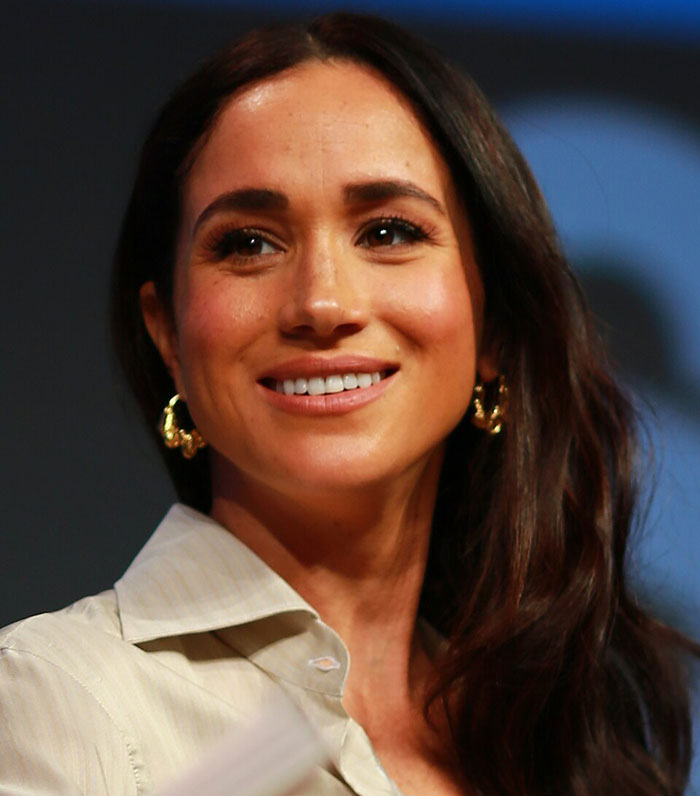 Meghan Markle smiling at an event, showcasing her natural look with loose hair and gold hoop earrings.