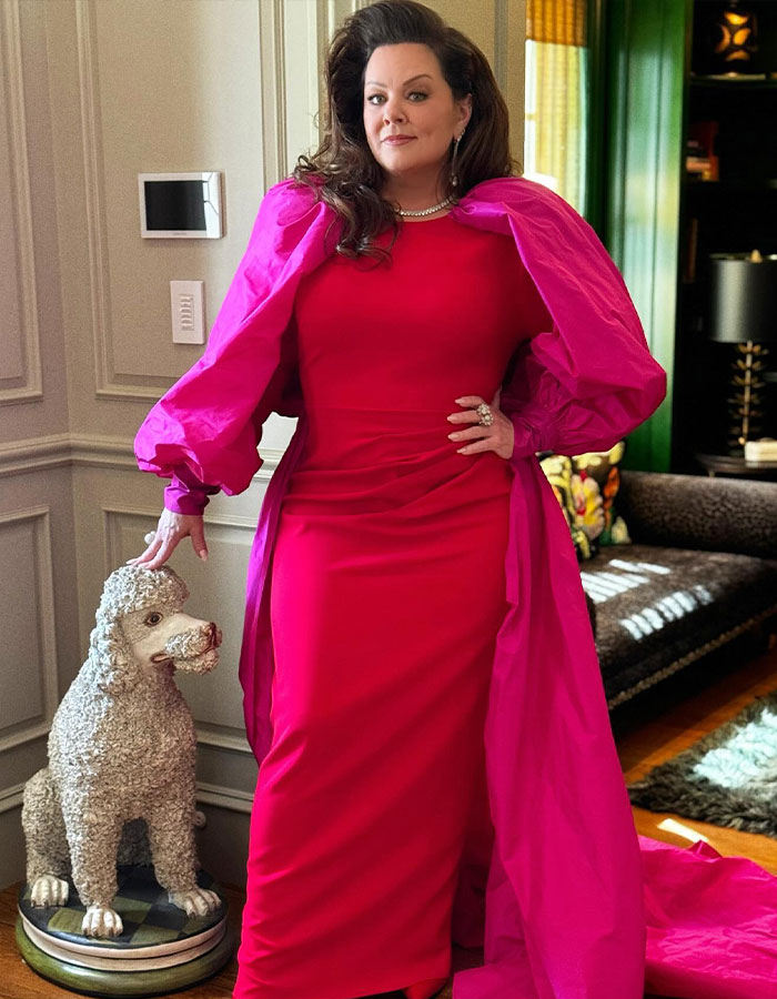 Melissa McCarthy in a bright pink gown posing indoors with a large statue of a poodle by her side.