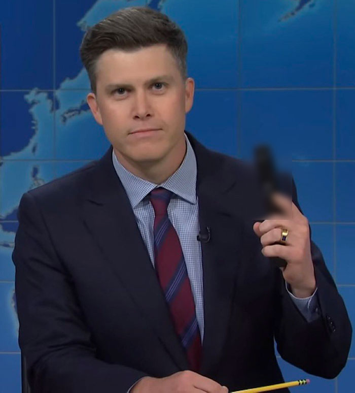 Man in a suit and tie holding a blurred object, appearing serious in a studio with a world map backdrop.