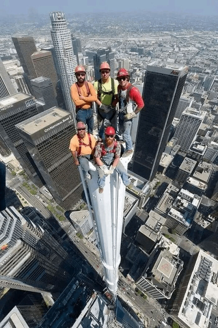 Five men wearing helmets and safety gear precariously perched atop a skyscraper, illustrating men doing dumb things.
