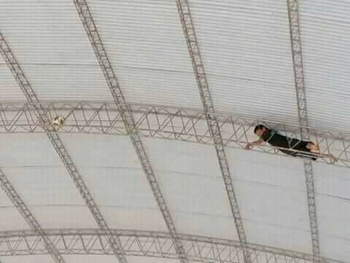 Man performing risky stunt hanging from metal structure under large roof, illustrating men doing dumb things concept.