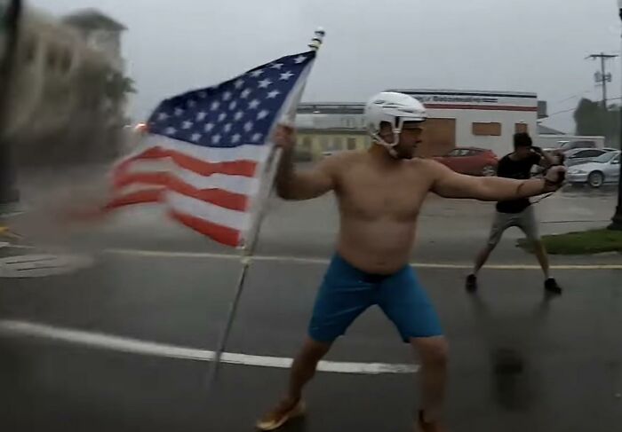 Man holding American flag wearing helmet and shorts on street, illustrating men doing dumb things outdoors.