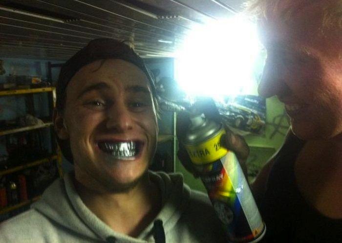 Young man with spray-painted teeth smiling while another person holds a can of spray paint in a cluttered garage, illustrating dumb things men do.