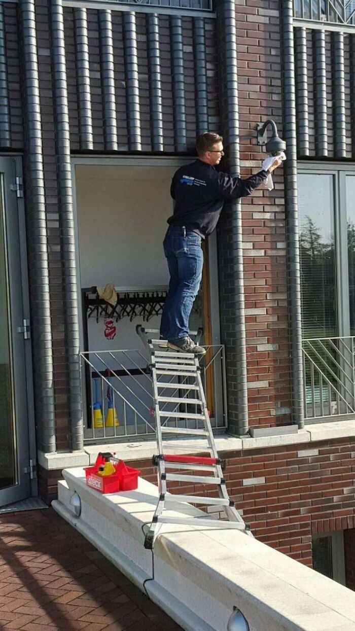Man doing dumb things stands unsafely on ladder on ledge repairing outdoor light fixture.