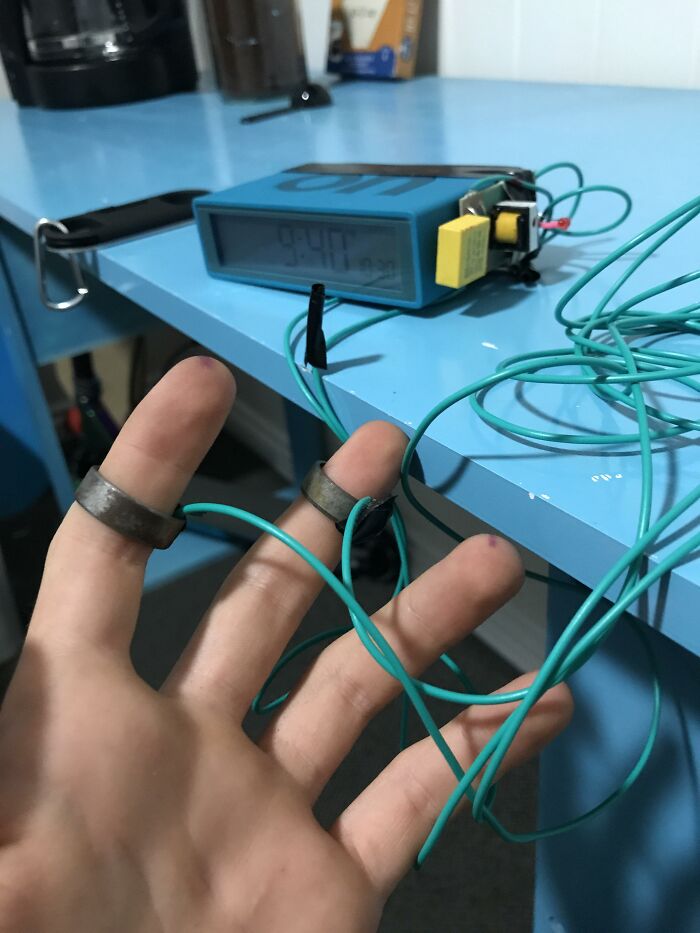 Hand with electrical wires wrapped around fingers near a timer device showing men doing dumb things humorously.