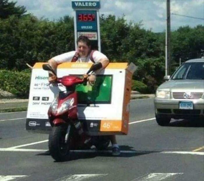Man riding a scooter while carrying a large TV box, illustrating men doing dumb things on the road.