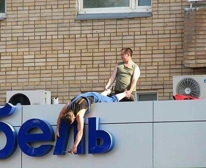 Two men doing dumb things on a rooftop, one hanging off a sign while the other holds his legs, illustrating risky behavior.