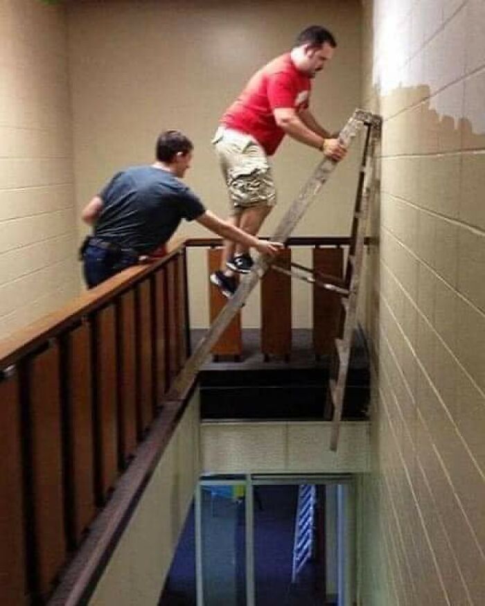 Two men dangerously using a ladder over a stairwell railing, illustrating men doing dumb things humorously.