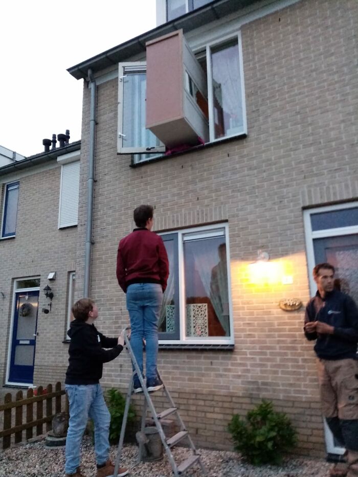 Two men attempting to lift a large cabinet through a second-story window showing men doing dumb things.