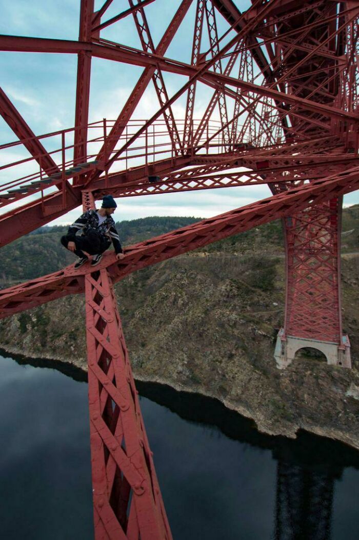 Man doing dumb things by climbing a high red bridge structure overlooking water and rocky terrain below.