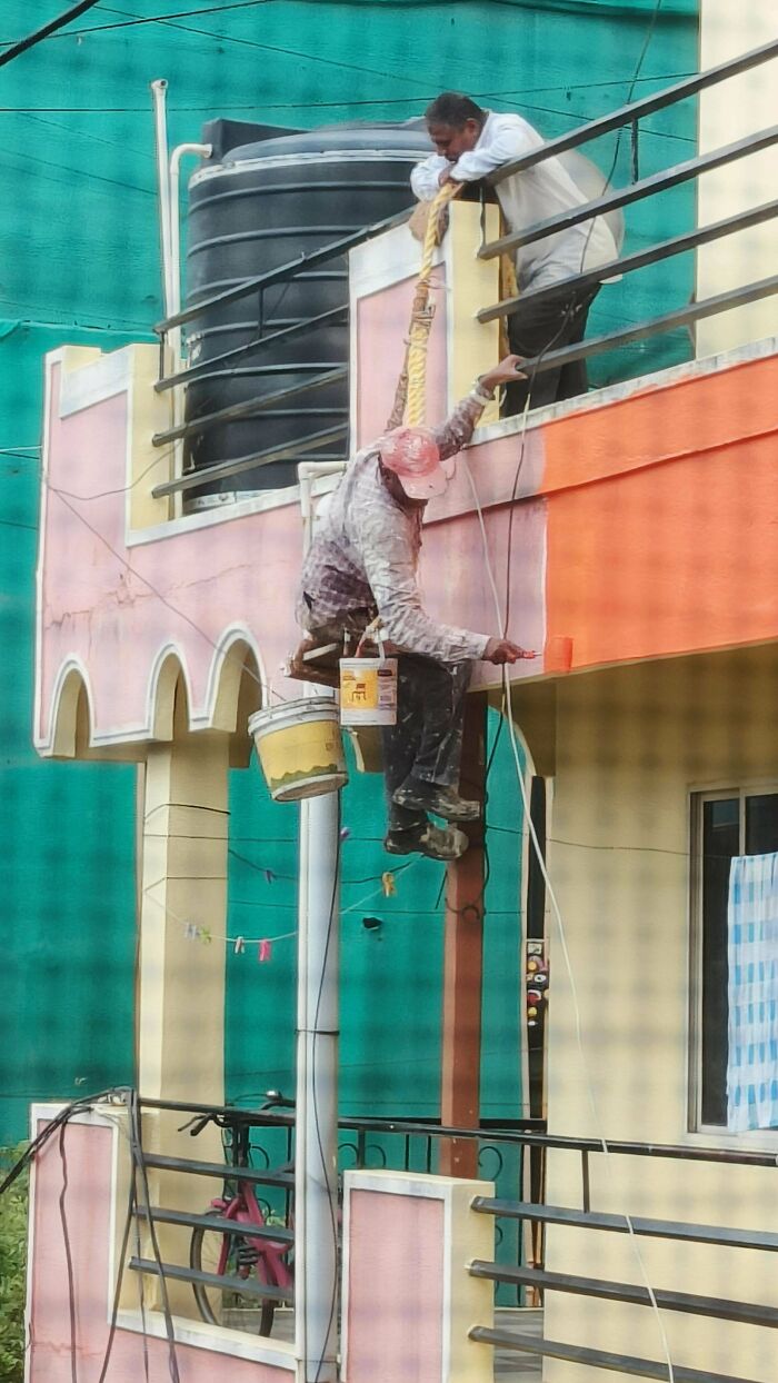 Man precariously painting a building while hanging from a rope, illustrating men doing dumb things outdoors.