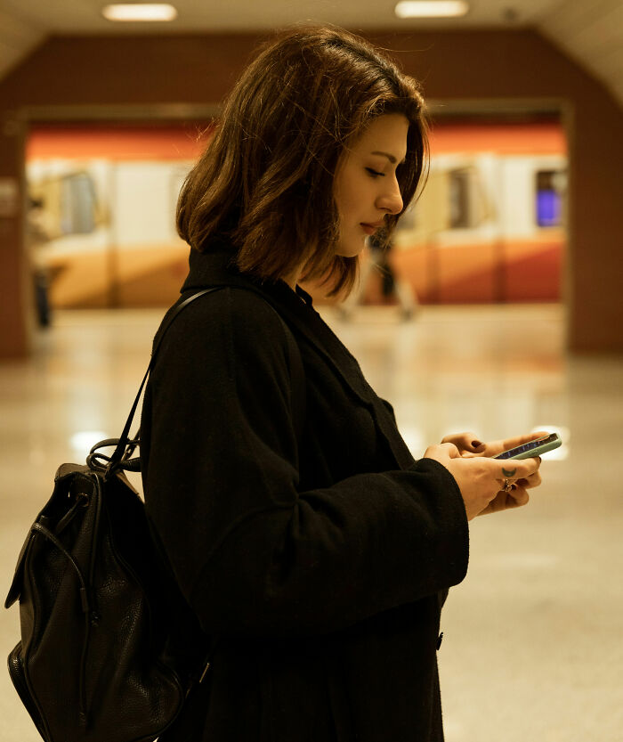 Young woman wearing black coat at subway station, focused on phone screen, illustrating postpartum OCD awareness.