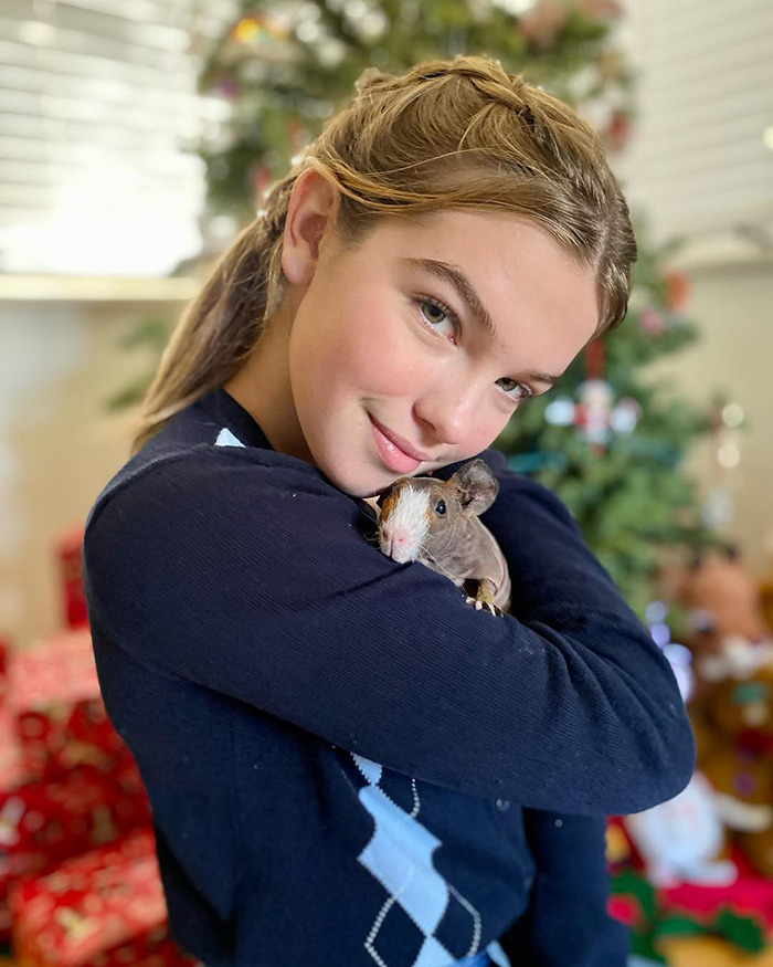 Teen girl and beloved child star from Good Luck Charlie hugging a guinea pig with a blurred holiday background.