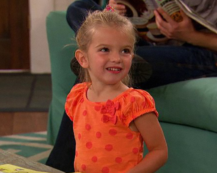 Young beloved child star from Good Luck Charlie smiling indoors, wearing an orange polka dot dress with short sleeves.