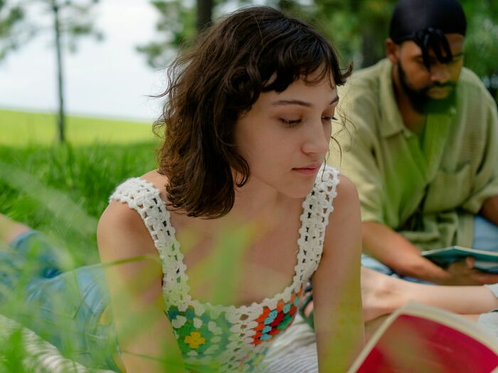 Young woman reading outdoors with a man in the background, illustrating challenges of modern dating experiences.