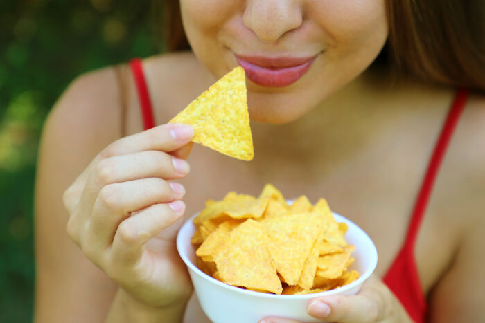 Young woman in a red top holding a bowl of chips and a single chip near her mouth, relaxed and enjoying a snack, completely legal total psychopath.