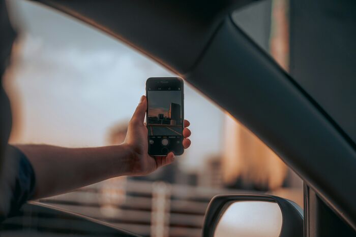 Person holding smartphone outside car window capturing photo of city skyline reflecting on screen, illustrating casual habits.